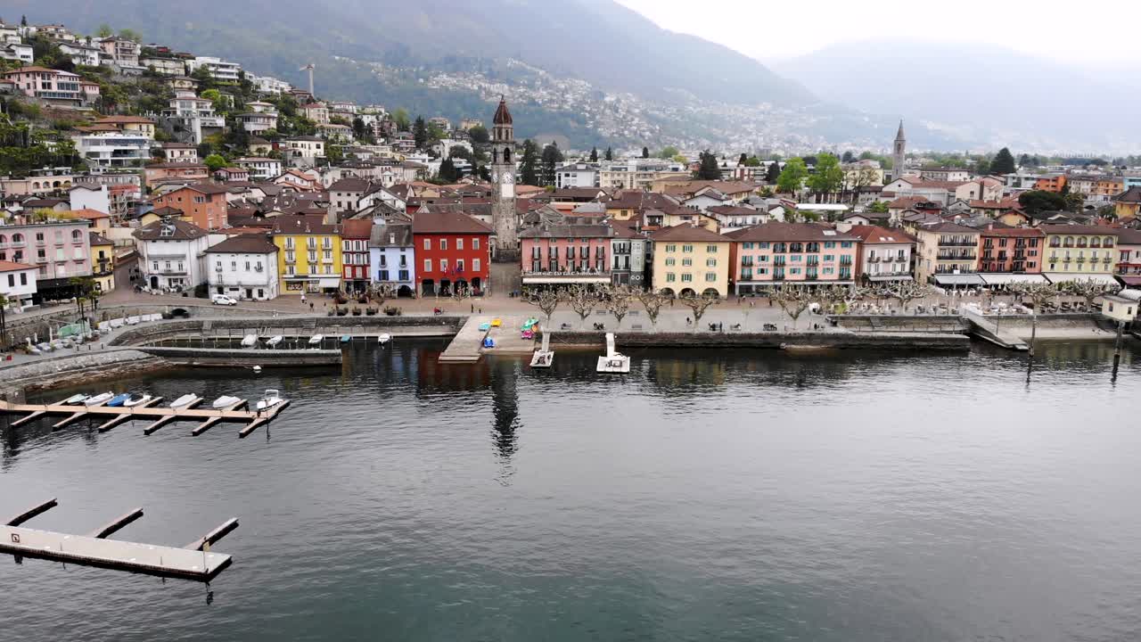 Aerial flyover along the shore of Lago Maggiore and promenade of Ascona in Ticino, Switzerland with a turn over the city rooftops revealing the lake and mountains behind the church tower