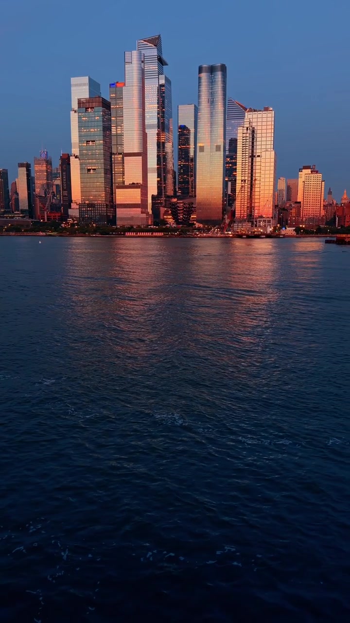 New York, USA, 7 June 2026 : Motorboat passing the illuminated Hudson Yards skyline, New York. A small white vessel cruises on dark blue river water in front of glowing sunset skyscrapers.