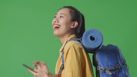 close up side view of asian female hiker with mountaineering backpack looking at smartphone then looking around while walking on green screen background in the studio