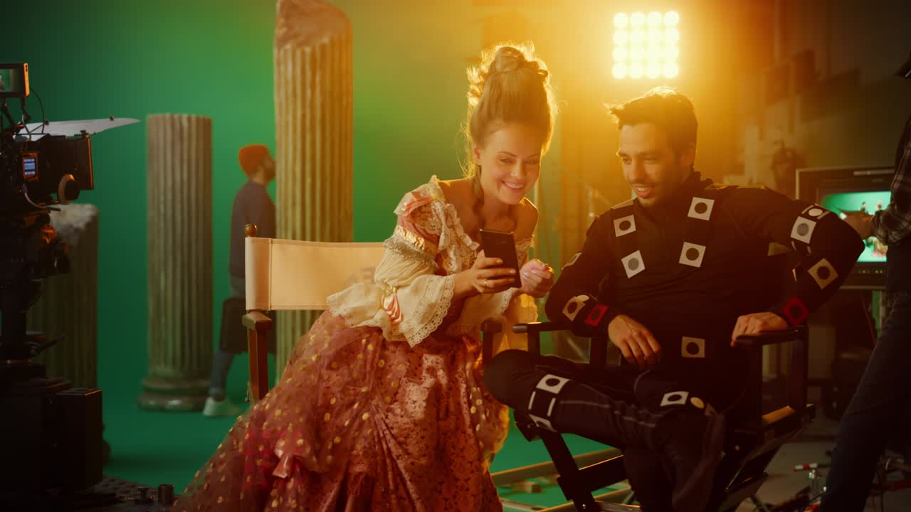 Beautiful Smiling Actress Wearing Renaissance Dress and Actor Wearing Motion Capture Suit Sitting on Chairs Share Social Media Posts via Smartphone. On Film Studio Period Costume Drama Film Set