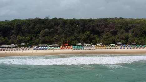 Left-trucking-aerial-drone-shot-of-the-famous-tropical-Madeiro-beach-near-Pipa,-Brazil-in-Rio-Grande-do-Norte-with-calm-waves-for-surfing,-colorful-umbrellas,-and-surrounded-by-large-exotic-cliffs