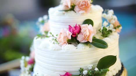 shot of a beautifully decorated floral wedding cake sitting on a pedestal right next to a pool during a summer wedding reception