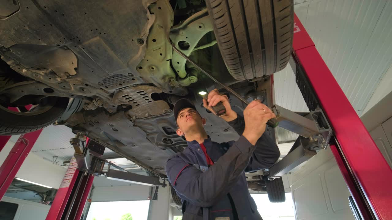joven mecánico de automóviles en una estación de servicio de reparación inspeccionando la rueda del automóvil y el detalle de la suspensión de un automóvil levantado. vista de abajo.