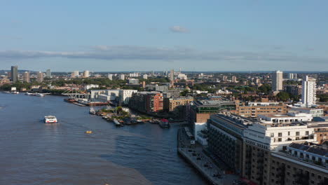 Forwards-fly-above-River-Thames-along-Butlers-Wharf-Pier.-Aerial-view-of-embankment-and-urban-neighbourhood-in-background.-London,-UK