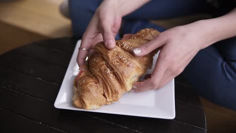 A-beautiful-brunette-in-casual-clothes-takes-a-large-croissant-sandwich-from-the-plate-and-bites-off-a-piece.-Eyes-are-closed-in-enjoyment.-Smiling.-Slow-motion