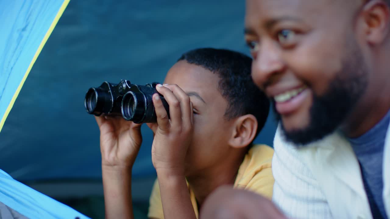 African American male adult leaning and pointing, guiding youth using binoculars at blue tent