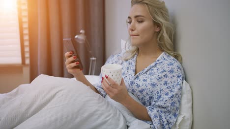 beautiful young woman browsing smartphone and holding mug of hot beverage sitting on bed