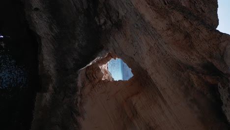 the drone is flying away from a hole in a rock cliff where a man is sitting on in vertical position in ibiza spain aerial footage 4k