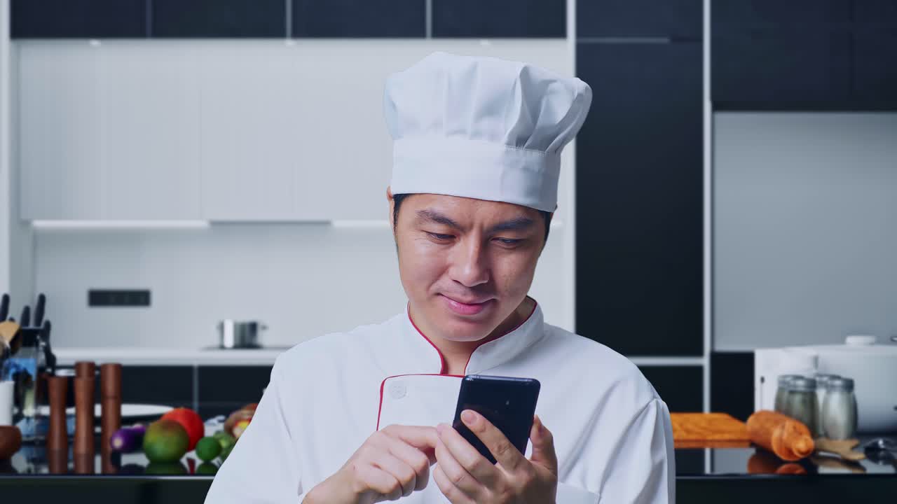 Gros plan sur un chef cuisinier asiatique souriant et montrant un smartphone dans sa main, debout dans sa cuisine.