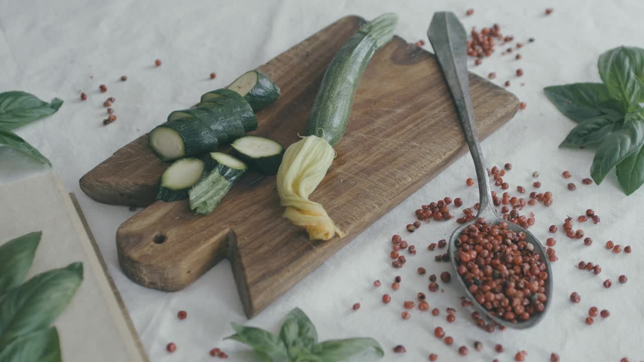 On a table there is a white tablecloth, an old spoon with red pepper, some basil and courgettes