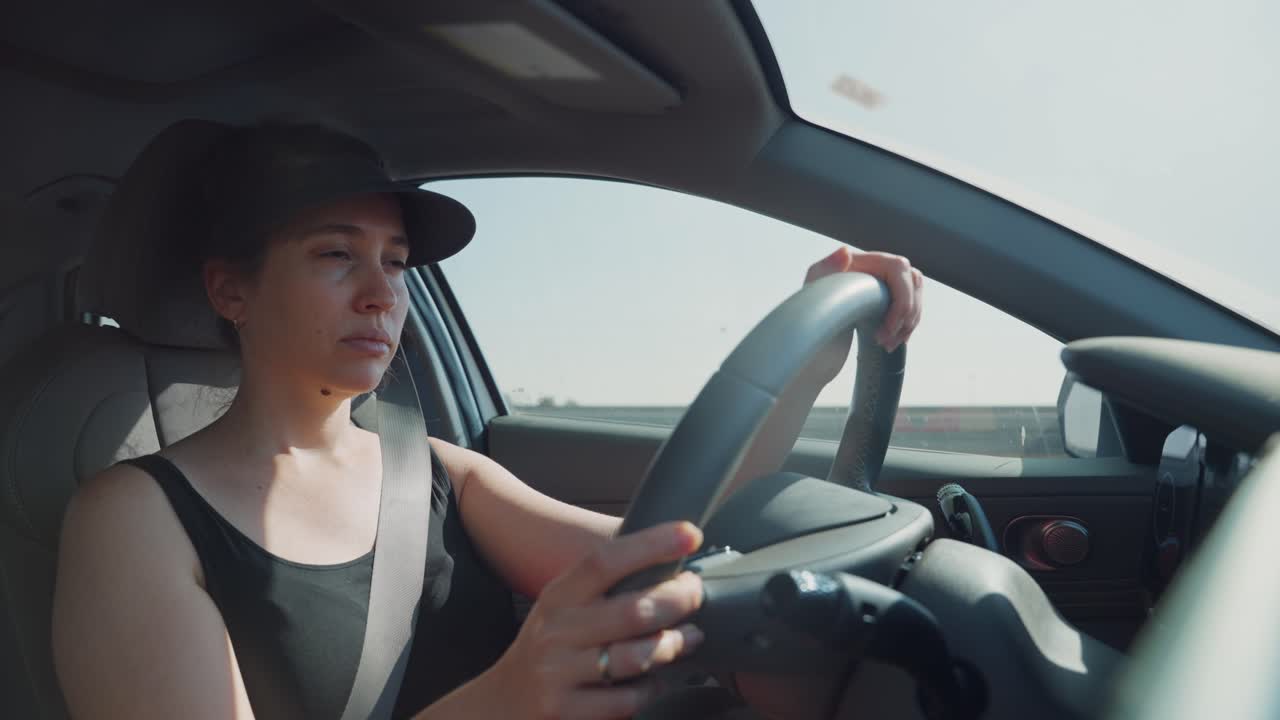Young woman driving modern hybrid car on highway on sunny hot day. Commuting to work or going on a road trip or vacation. Tired and bored she looks on road ahead. POV from dashboard cam as she drives
