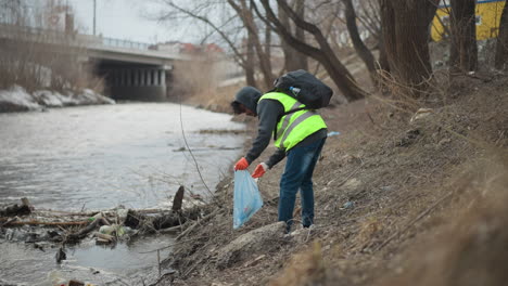 volunteer wearing reflective vest and gloves collects trash along polluted riverbank near bridge, placing waste into plastic bag during environmental cleanup to protect nature and improve urban waterway