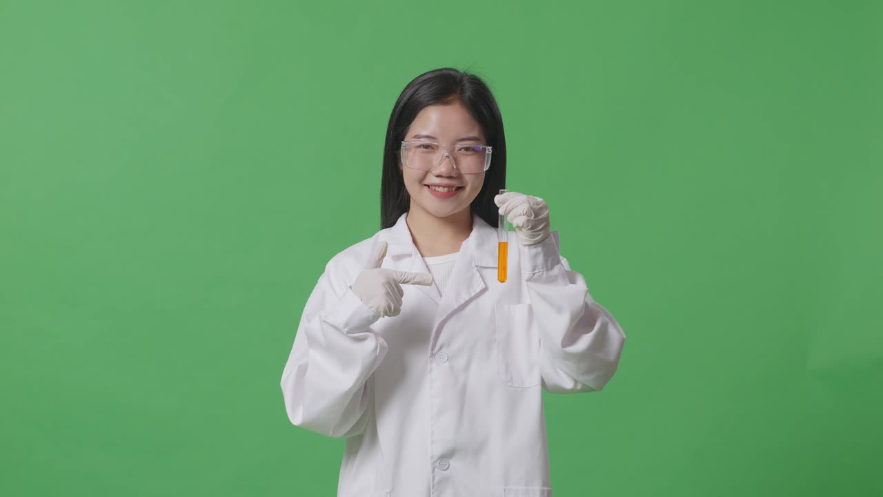 아시아 여성 과학자가 미소 짓고 실험실의 녹색 스크린 배경에 서서 손에 있는 시험관에 있는 오렌지색 액체를 가리키고 있습니다.