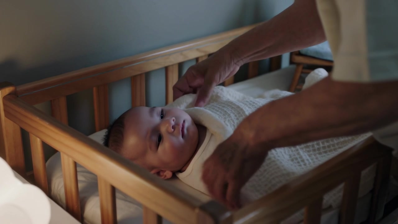 Approaching caregiver lifting infant into wooden crib under cloud lamp, swaddling blanket for sleep
