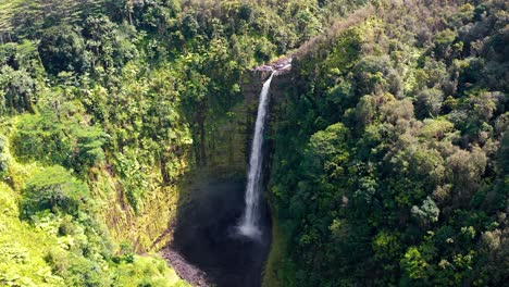 a towering waterfall streams down a vertical rainforest cliff into a dark volcanic basin, captured from above, showcasing the immense scale and untamed tropical wilderness