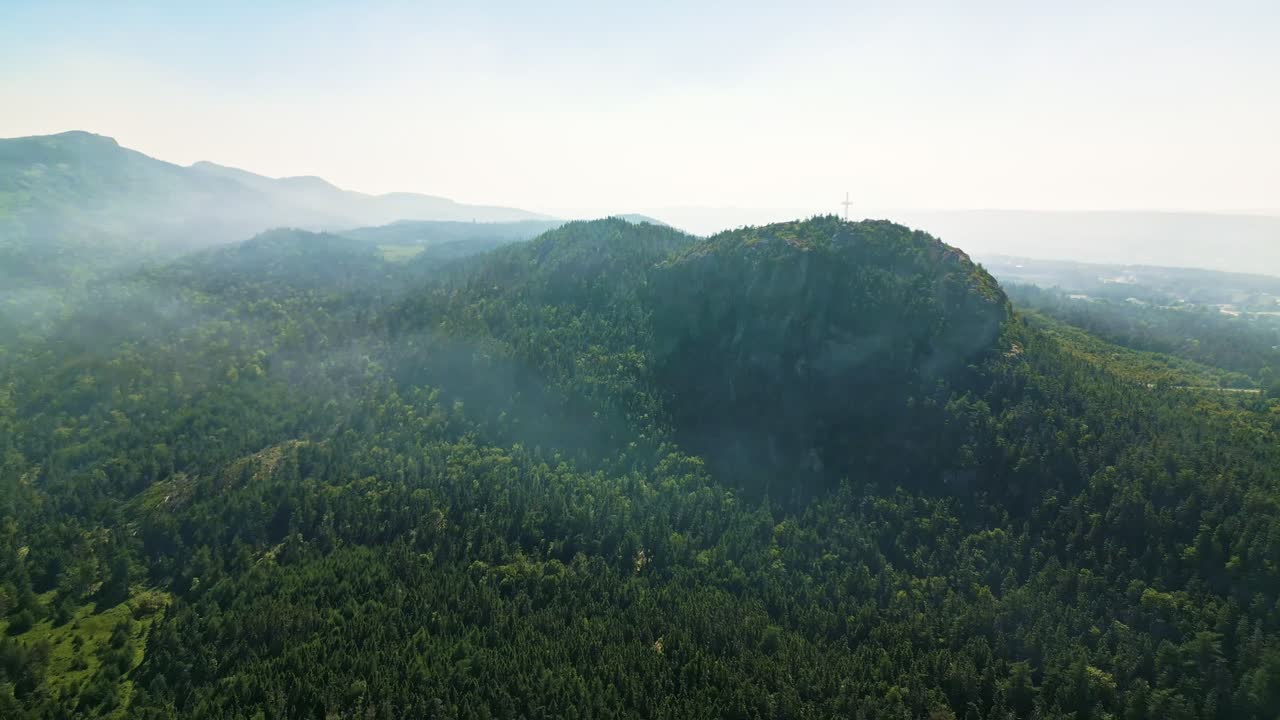 A drone glides over Holyrood's dark green hills and valleys while a generating station's smoke plume drifts across the sky, blending with the hazy horizon