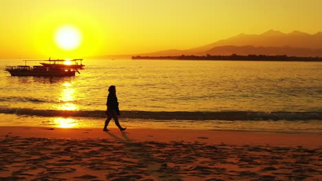 woman-walking-along-the-beach-at-sunset-in-French-Polynesia,-boats-floating-on-the-calm-sea-golden-colored-sun-and-horizon