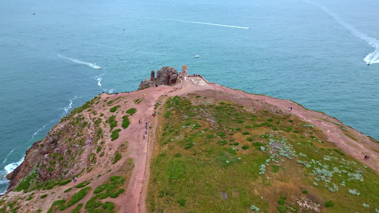 People walking along paths on rugged Cap Fréhel headland, old tower, boats on sea, Brittany, France. Aerial forward