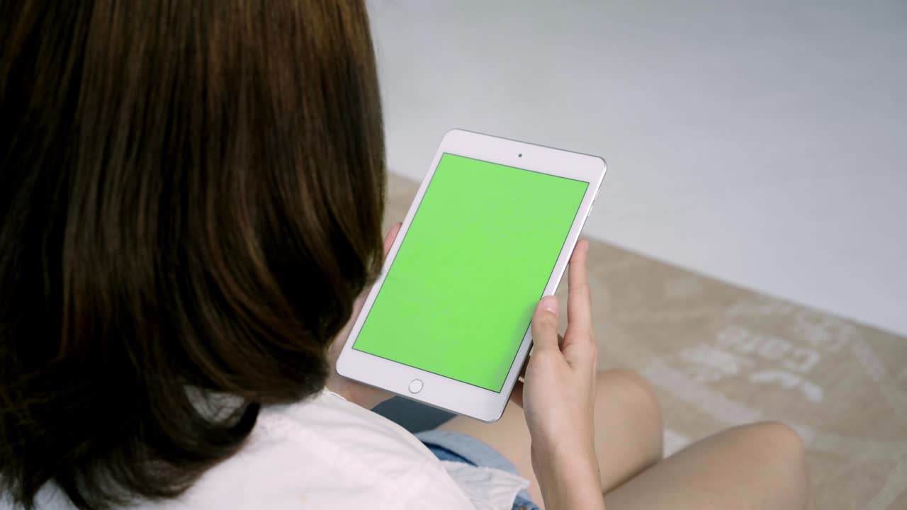 mujer joven asiática usando un dispositivo de tableta negra con pantalla verde. mujer asiática sosteniendo una tableta, desplazando páginas mientras está sentada en el sofá en la sala de estar. clave de croma.