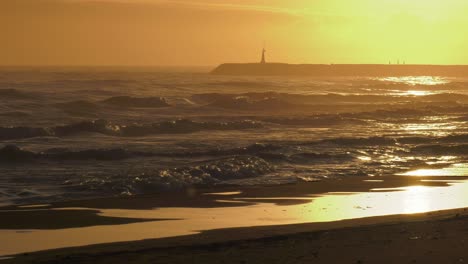 Rough-seas-at-sunrise,-waves-rolling-onto-sandy-beach-with-harbor-wall-in-background