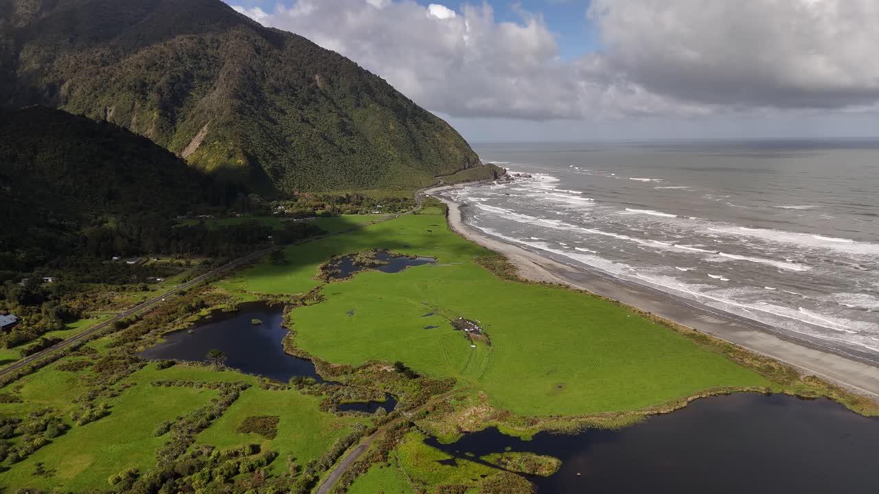 Barrytown sulla costa occidentale dell'Isola del Sud della Nuova Zelanda, che mostra zone umide, campi costieri verdi e onde oceaniche sotto ripide montagne, sotto nuvole luminose e cielo aperto. Veduta aerea dall'alto.