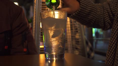 person pouring a drink over a glass with ice on a bar table with a stove with a fire flame in the background