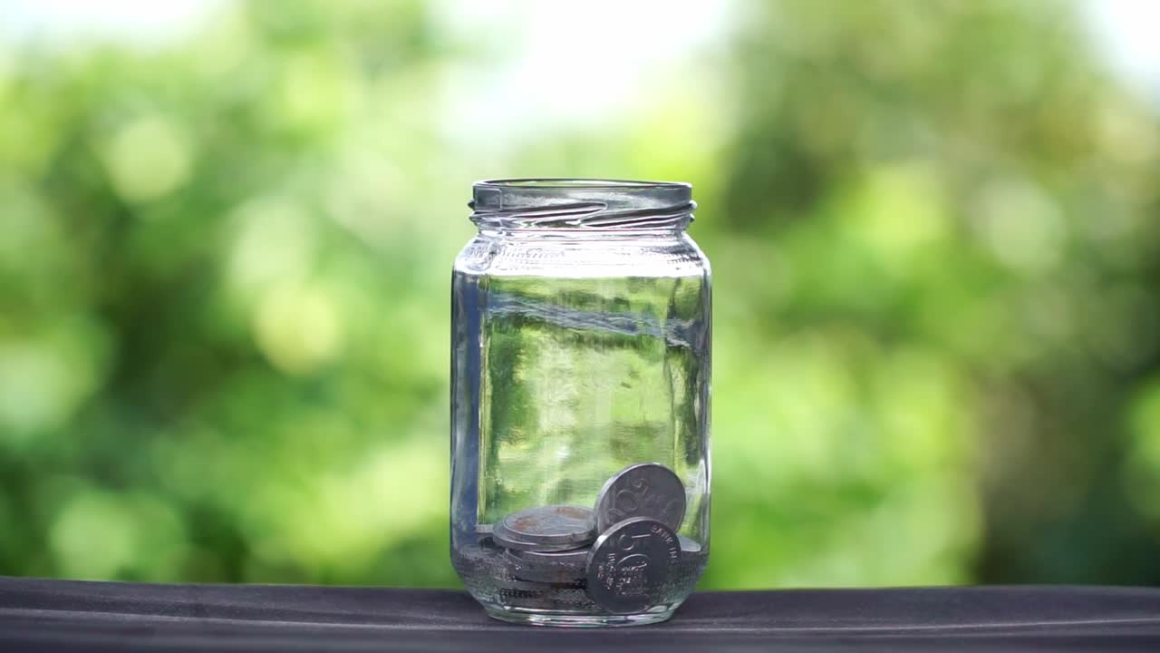 自然の緑を背景にガラス瓶にお金を入れる、貯蓄のコンセプト。