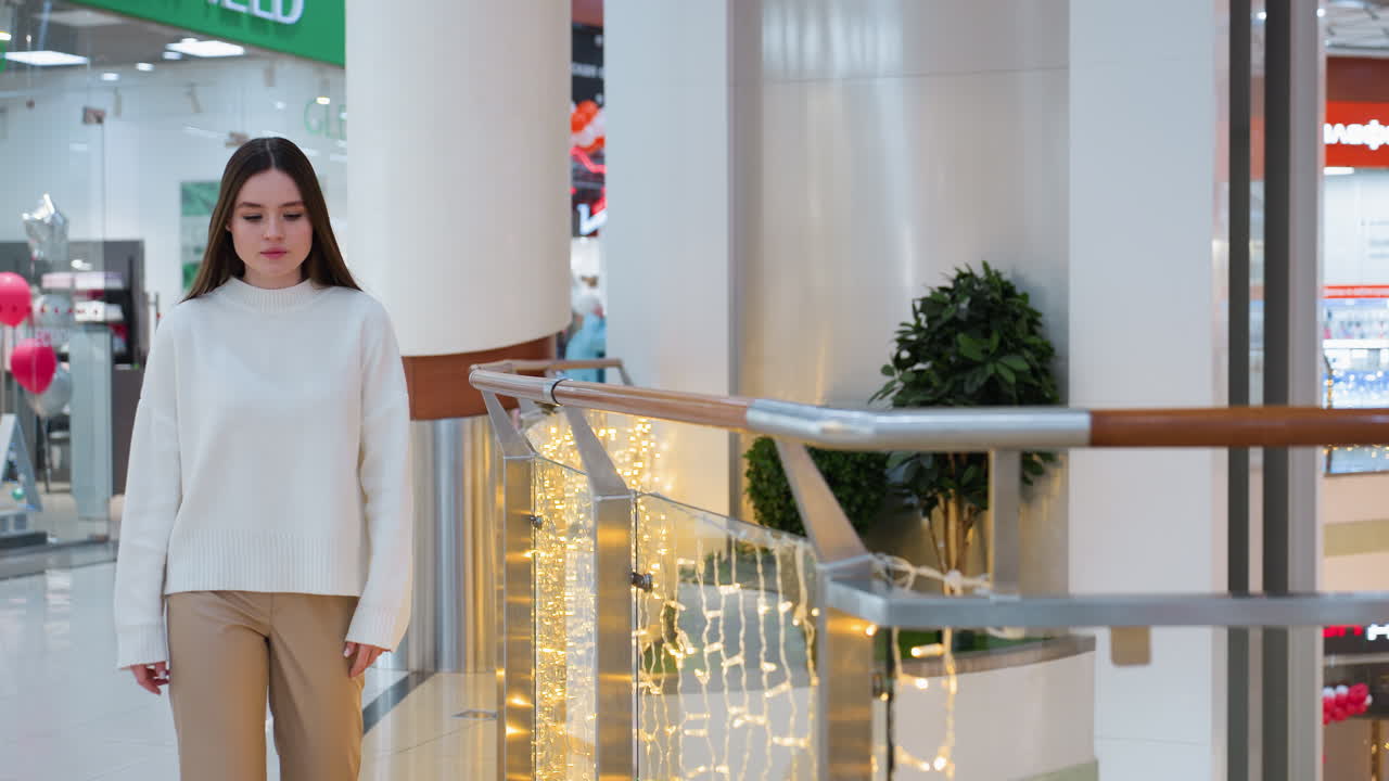 Elegant woman in white sweater walks through beautifully decorated shopping mall with festive lights, moves towards railing while other shoppers browse in background
