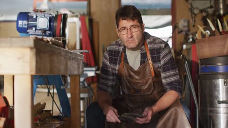 caucasian male knife maker sitting in workshop, looking at camera and smiling