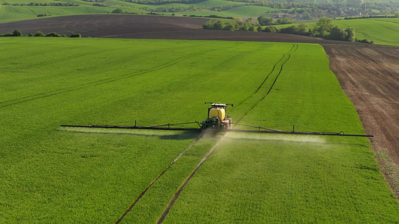 Vista aerea con drone di un'irroratrice agricola con ampi bracci che irrora campi di grano verde con nebbia di spruzzo visibile e colline agricole ondulate sullo sfondo, Moravia, Repubblica Ceca.