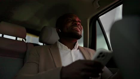 a happy man with black skin with a beard in a brown suit sits on the passenger seat and listens to music in white wireless headphones during his vacation in a modern car interior in the city