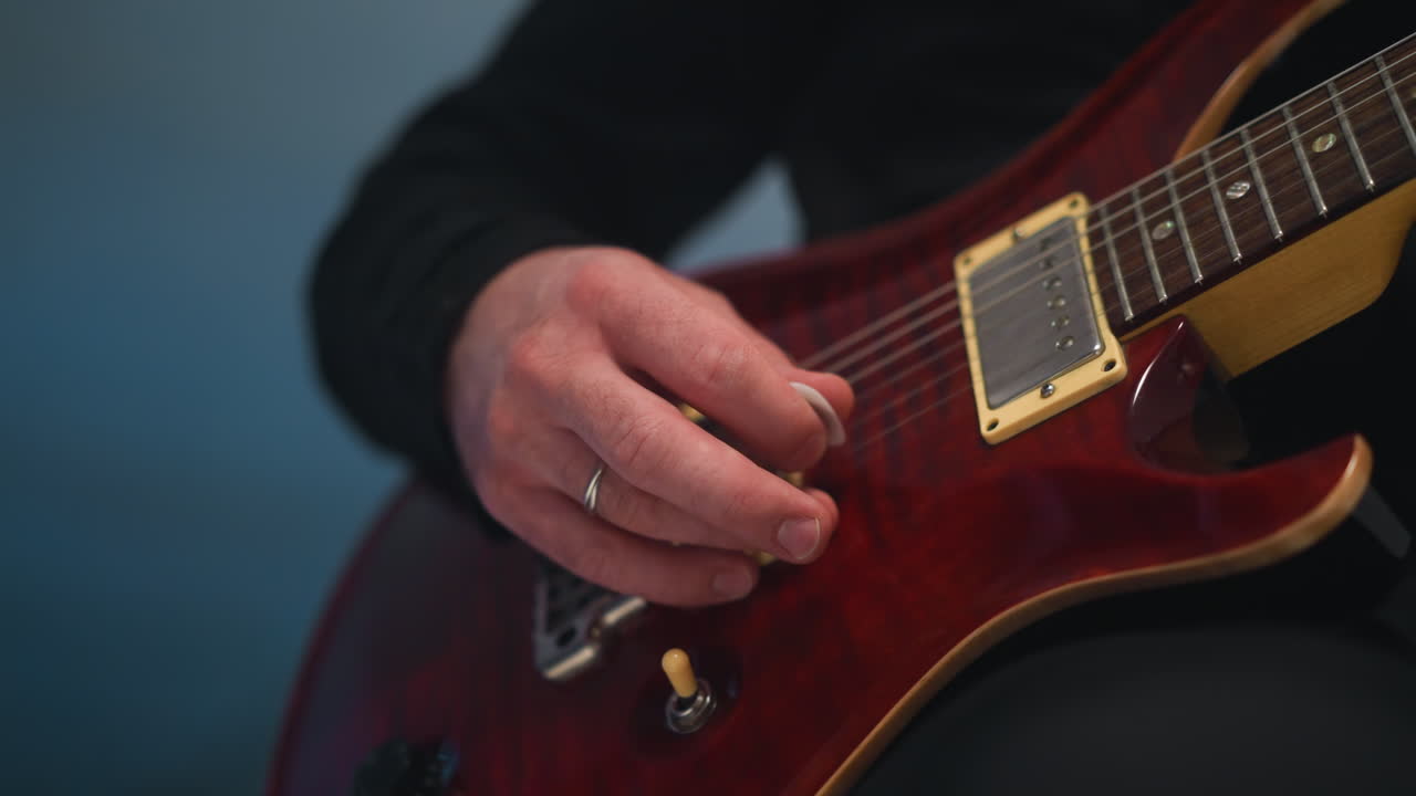 vista de cerca de la mano de un guitarrista tocando una guitarra roja contra un fondo azul, destacando los detalles intrincados del instrumento y los dedos hábiles del músico
