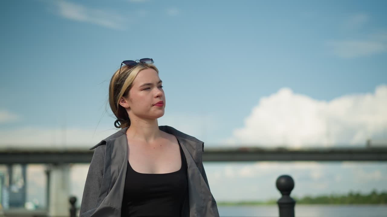 mujer en una camiseta negra y gris abierta camina cerca de un puente, mirando a la distancia, el fondo presenta un borrón de coches que pasan por el puente bajo un cielo azul claro