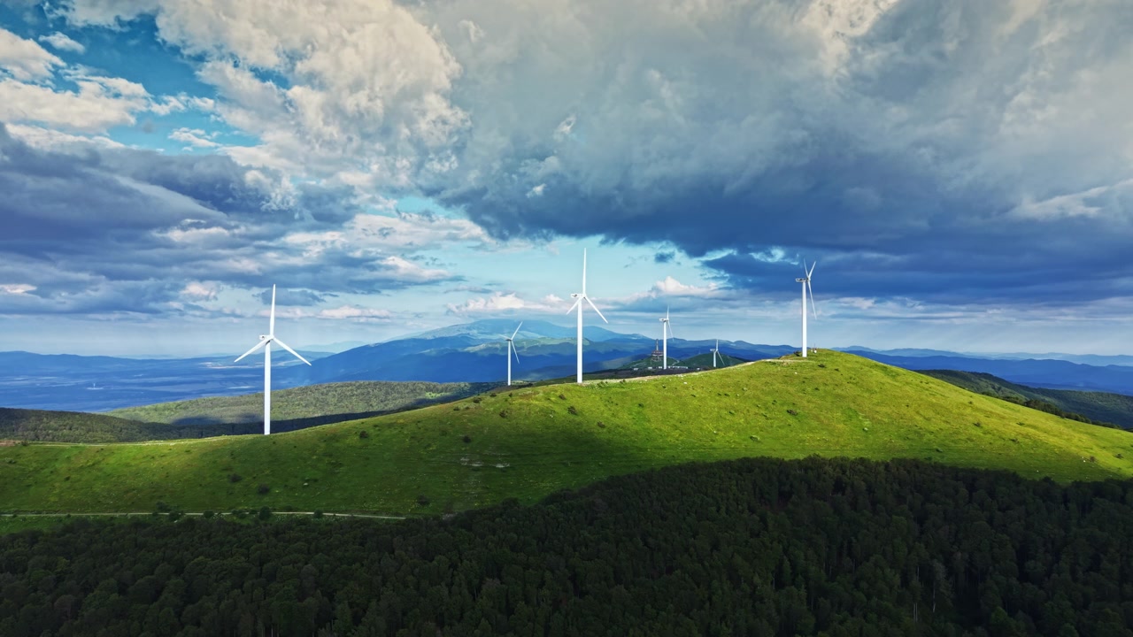 Le turbine eoliche si ergono su una collina verde sotto un cielo nuvoloso con montagne in lontananza durante il giorno