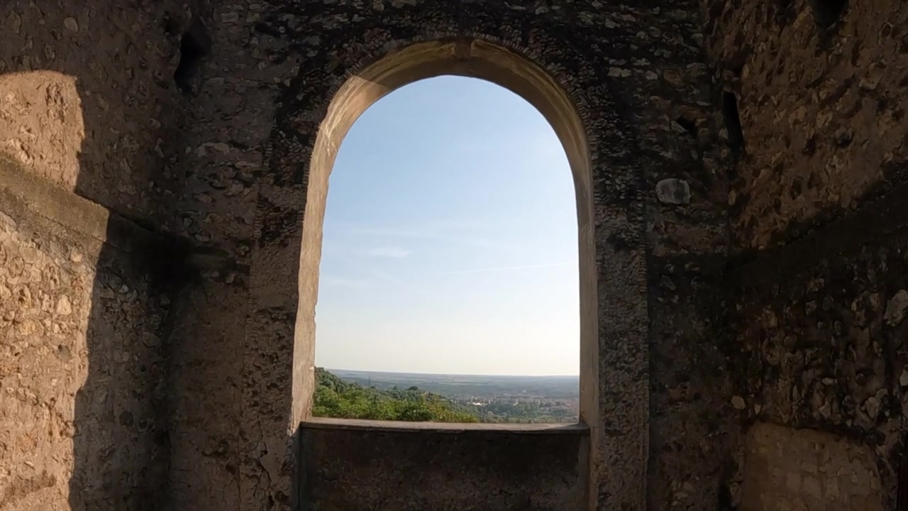 Plan frontal en mouvement d'une tour de château médiéval en pierre à Tivoli, avec des arbres verts luxuriants, un portail voûté et un horizon lointain.