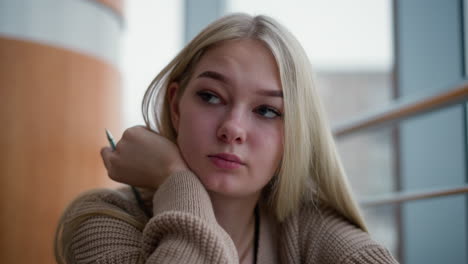 young lady seated, resting chin on hand with pen in hand, looking tired and exhausted while glancing at busy urban setting through window, soft blur of city life outside