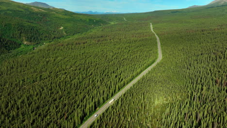 a bus and a car going by the long highway crossing the valley covered with vast evergreen woods. flight over the wilderness in alaska, usa on sunny day