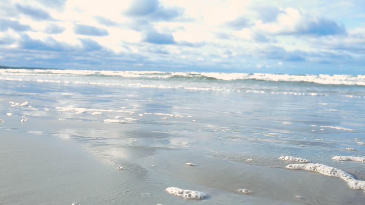 Close up shot of a sandy beach shoreline covered with ocean sea water foam and bubbles and waves in the background moving in slow motion during day time in Vääna Jõesuu beach, with dark clouds in sky.