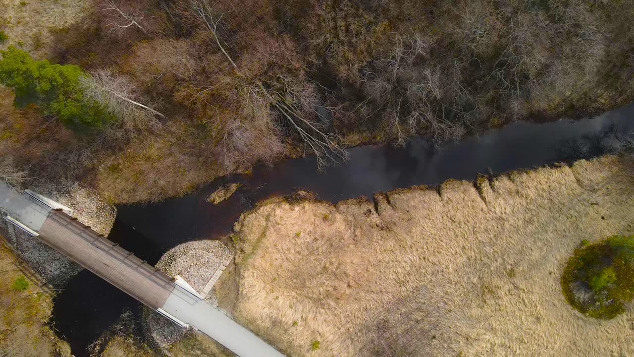 Top down aerial drone view decending over a wooden brown bridge that is crossing a narrow black and dark colored Pääsküla river during sunny spring or autumn day. Brown overgrown grass on river sides