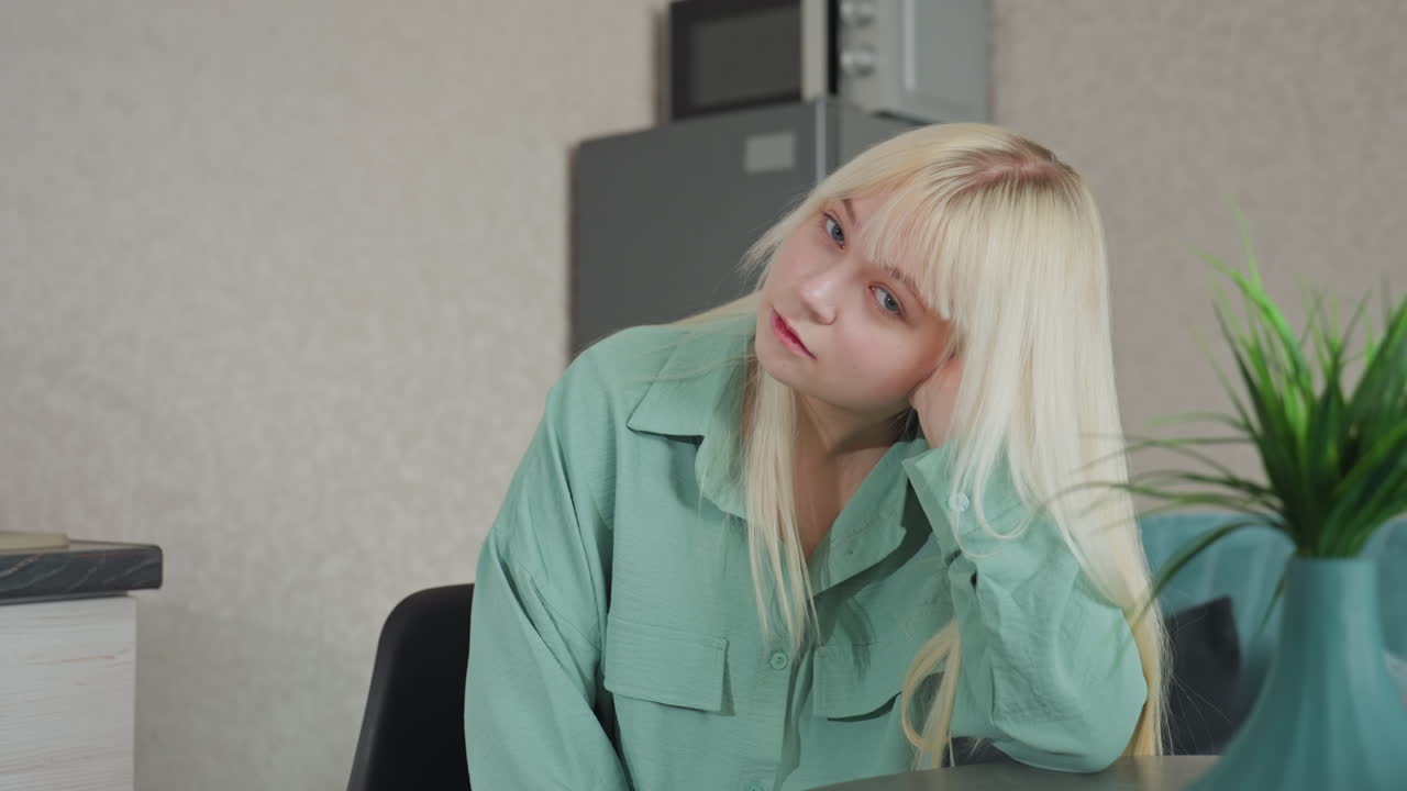young woman in green shirt resting head on hand thoughtfully while sitting at kitchen table with decorative plant on table and fridge, microwave in background in modern apartment