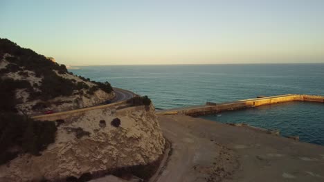 Drone-view-of-a-coastal-road-winding-along-Costa-Garraf-cliffs-at-sunset-in-Barcelona,-Spain