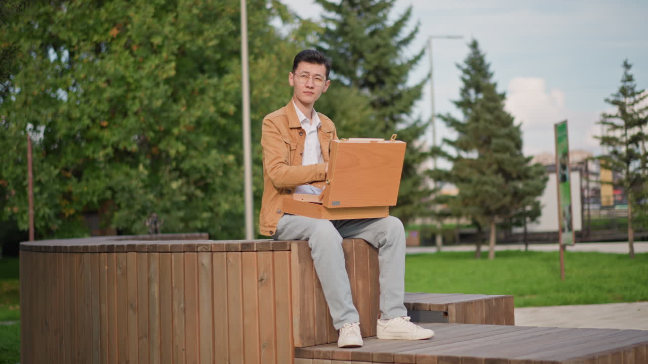 artist sketching outdoors on bench with wooden portfolio, young man concentrates on urban sketch of nearby trees and pastel buildings, casual jacket and sneakers, soft natural light and calm park