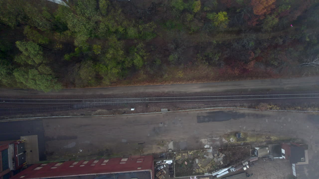 Aerial drone view of the end of a long freight train passing through an industrial area bordered by trees and scattered debris