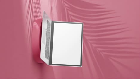 portátil plateado elegante en un pedestal redondo, pantalla vacía con sombras de palma sobre fondo rojo, vertical