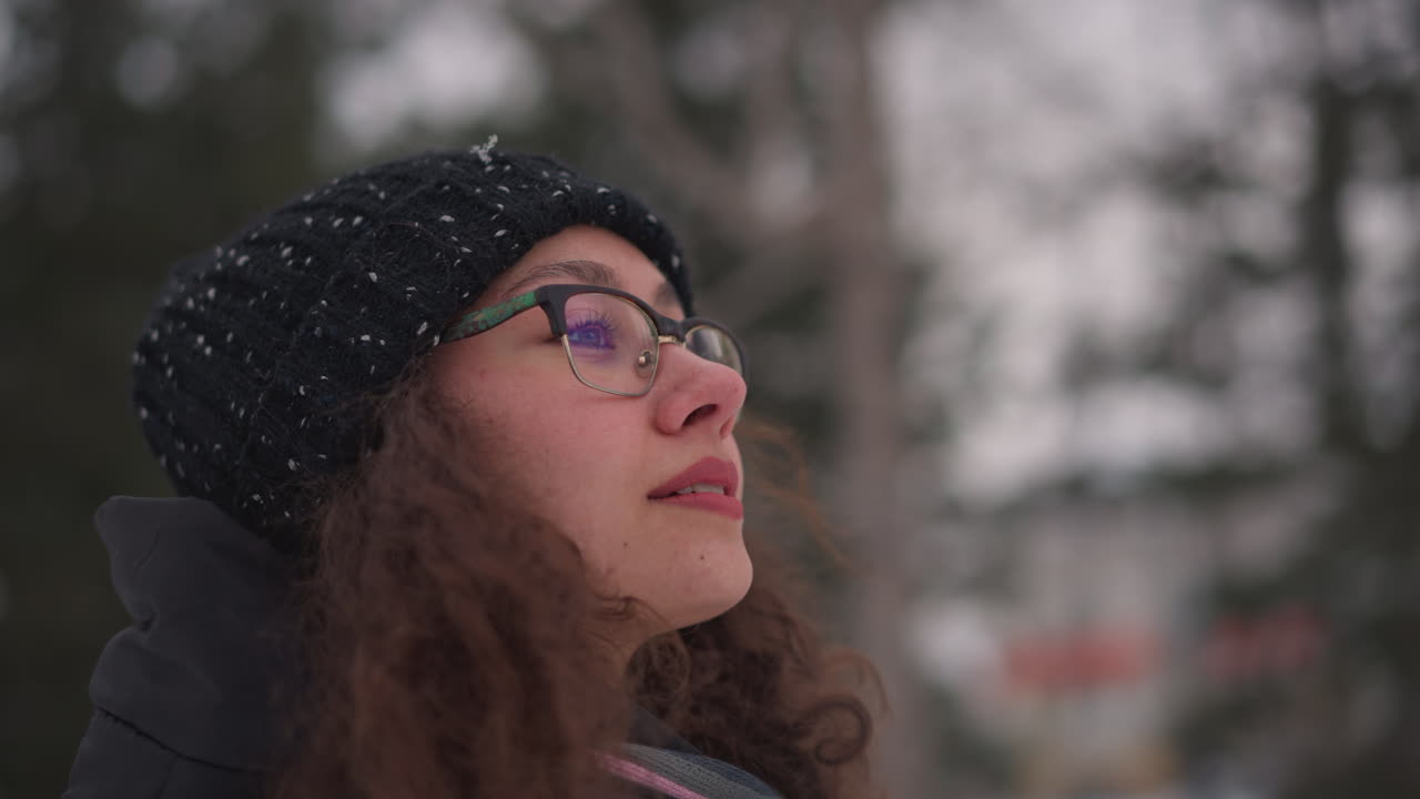 Female wearing glasses and black knitted hat outdoors in snowy environment close up side face view with curly hair looking thoughtful winter season background blurred frosty forest atmosphere