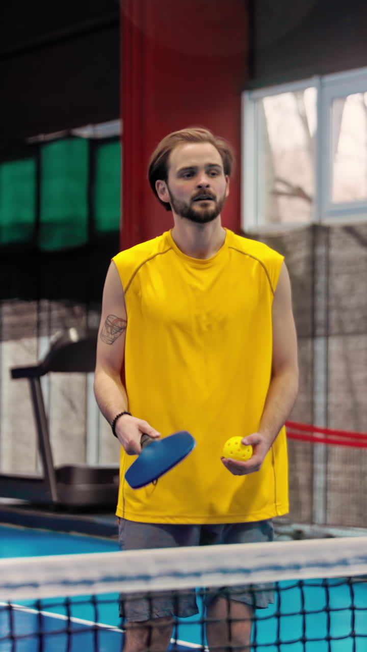 Man in yellow shirt playing pickleball on a blue, inside court. Vertical