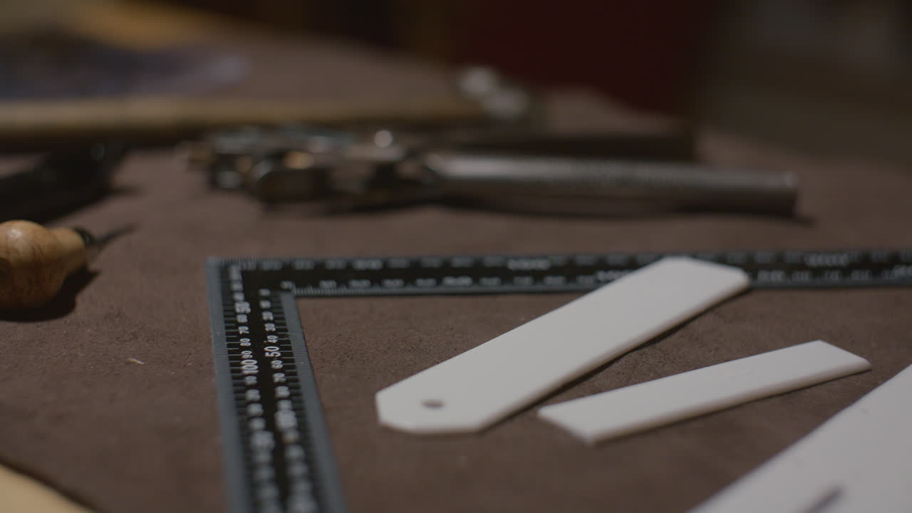 Leather crafting tools, including a ruler, spacers, and other equipment, placed on a brown leather work surface in a workshop setting.