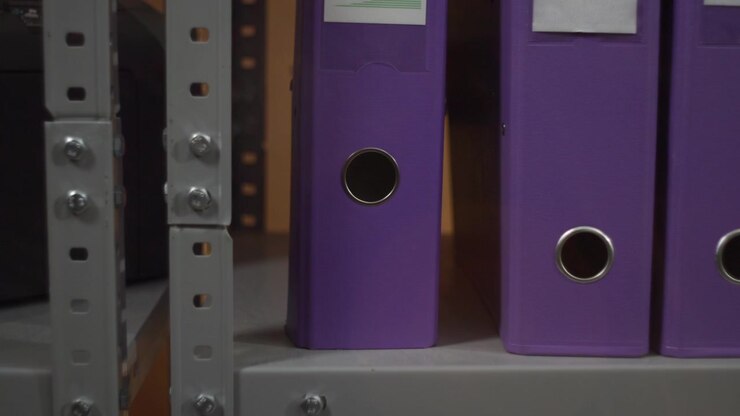 Primo piano di raccoglitori ad anelli viola in piedi su uno scaffale con un bordo grigio, illuminati da una luce interna soffusa. La disposizione ordinata suggerisce un archivio per ufficio.