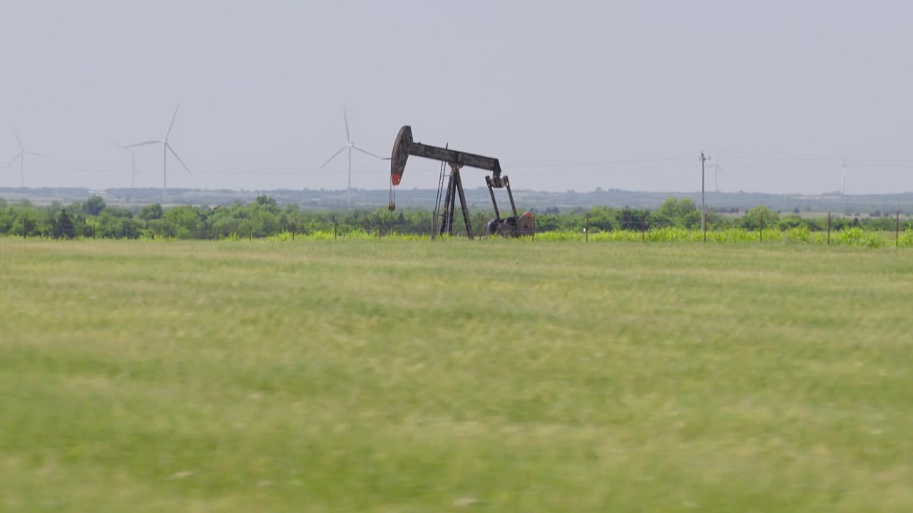 Drone 4K of a green field with an oil pumpjack and wind turbines in the background.