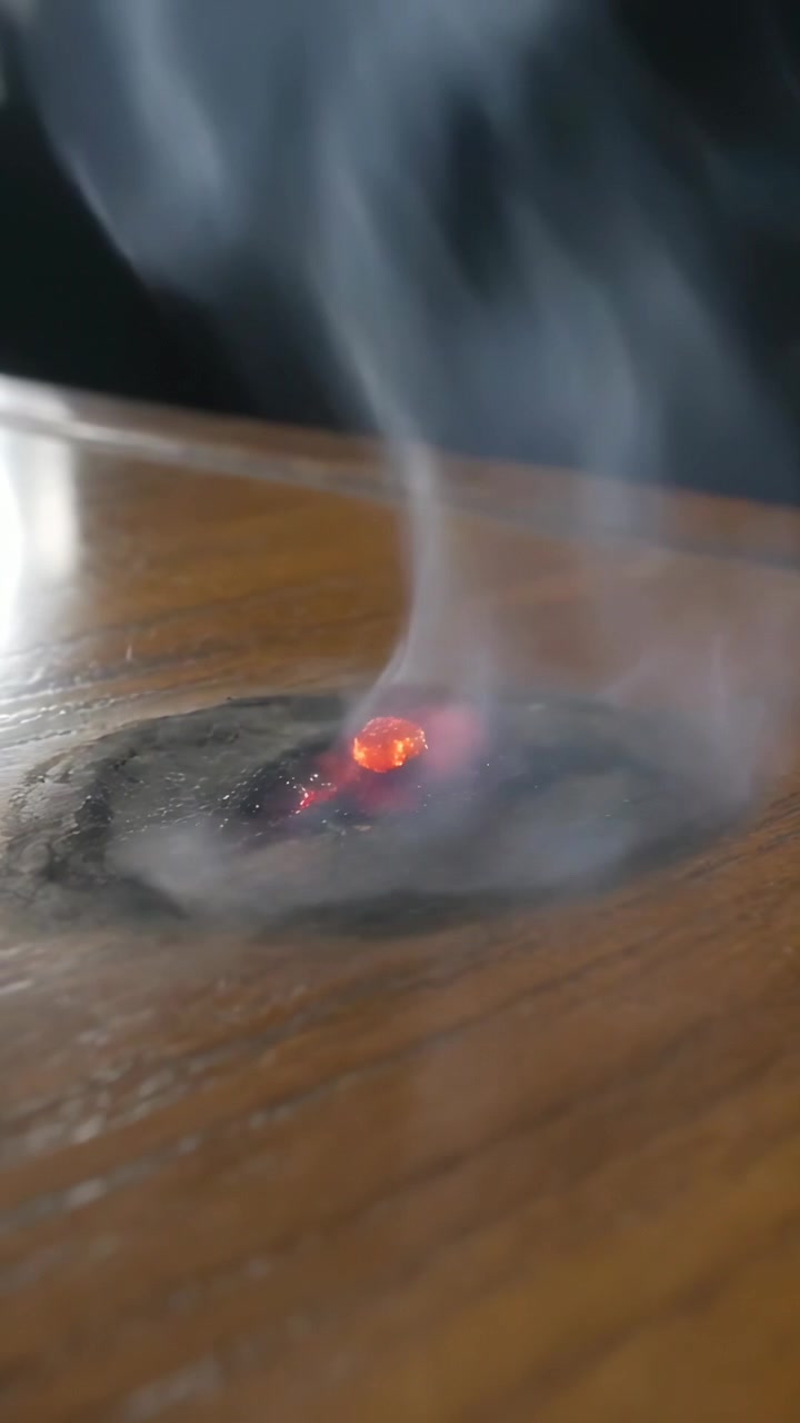 Vertical video: Landing ember smoldering on polished wooden table, producing smoke and charred ring